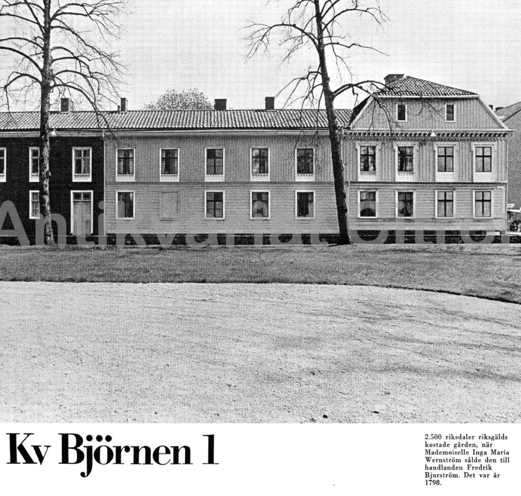 "Fasad till Björnen 3 A i Jönköping, en tvåvånings träbyggnad med klassiskt profilerade fönsteromfattningar, liggande gråmålad panel och takfot med enkel listverkan. Huvudentrén har en äldre spegeldörr med överstycke, flankerad av vertikalt placerade ljusinsläpp. Byggnaden visar tydlig påverkan från 1800-talets urbana trähusmiljöer."